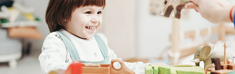 Ein kleines Kind mit kurzen braunen Haaren lächelt, während es an einem Tisch mit Holztieren spielt. Eine erwachsene Hand hält ein Spielzeugtier vor dem Kind hoch. Der Hintergrund ist leicht verschwommen.