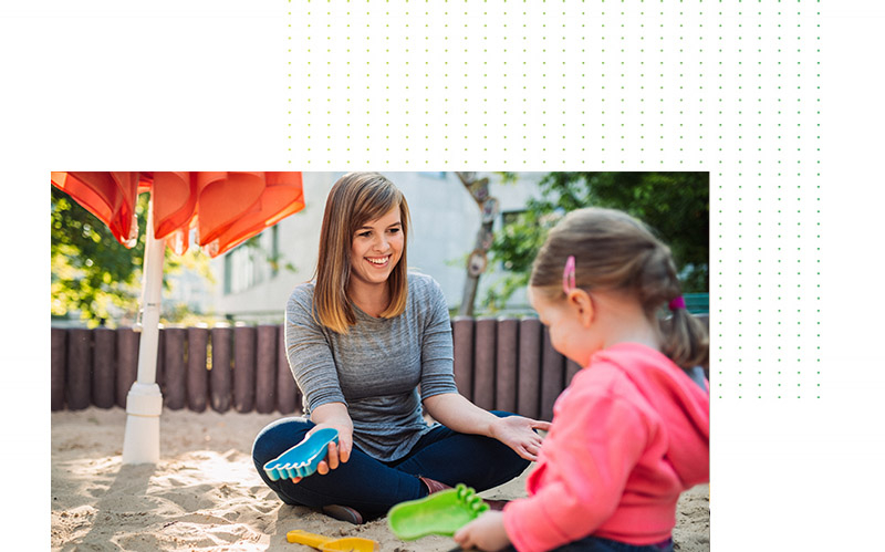A woman is sitting in a sandpit with a little girl. They are smiling at each other and playing with colorful sand toys.