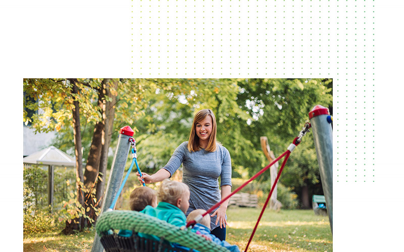 A woman pushes three small children in a large nest swing on a playground. Trees and play equipment can be seen in the background.