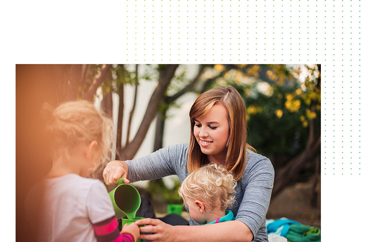 A woman sits outside with two small children and pours water from a play jug. The children watch curiously.
