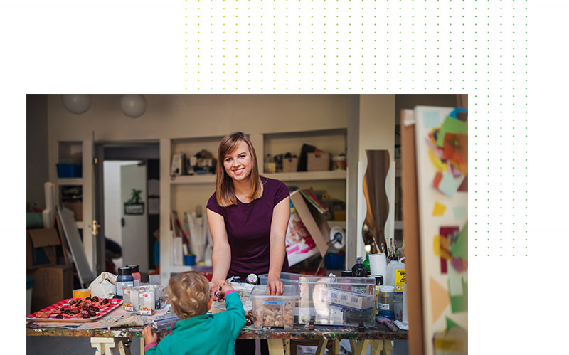 A woman stands at a craft table and talks to a small child. The room is full of materials and looks like a creative studio.