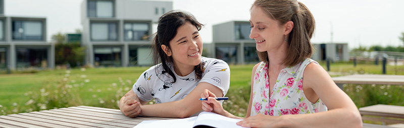 Zwei Studentinnen sitzen an einem Tisch im Freien vor einem Wohnheim. Eine zeigt der anderen etwas in einem offenen Buch.