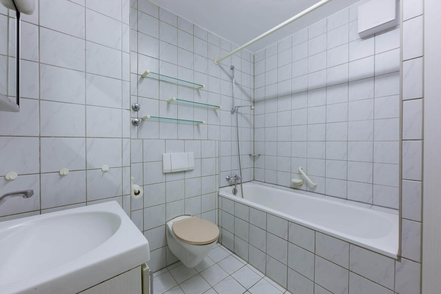 Bathroom with bathtub and shower curtain, wall-mounted WC and washbasin; fully tiled in light tones with glass shelves on the wall.