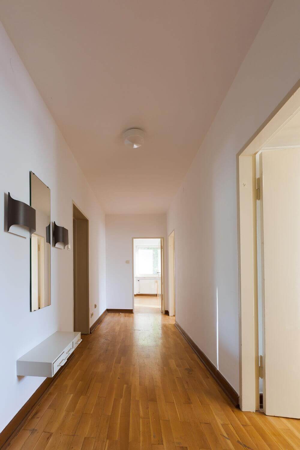 Long hallway with wooden floor, white walls, wall mirrors and several room doors; at the end of the hallway a window with natural light.