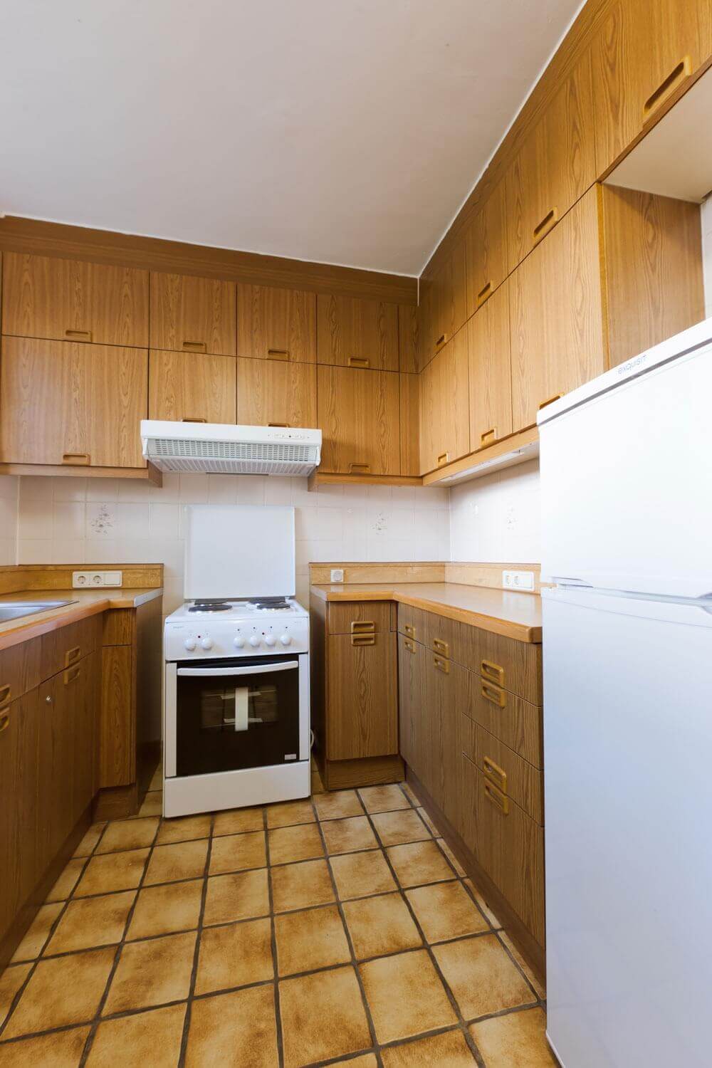 Shared kitchen with brown wooden cupboards, electric stove with extractor hood, fridge and retro-style tiled floor.