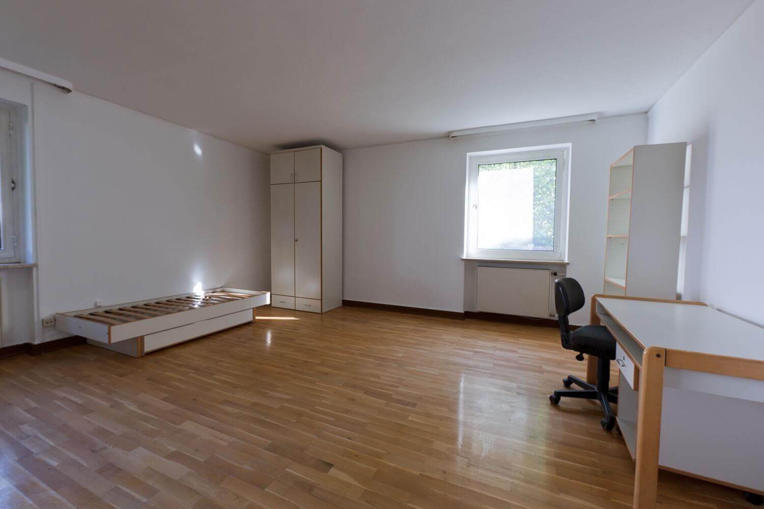 Spacious, simply furnished single room with parquet floor, desk, shelf and bed frame without mattress; daylight through a window on the right wall.