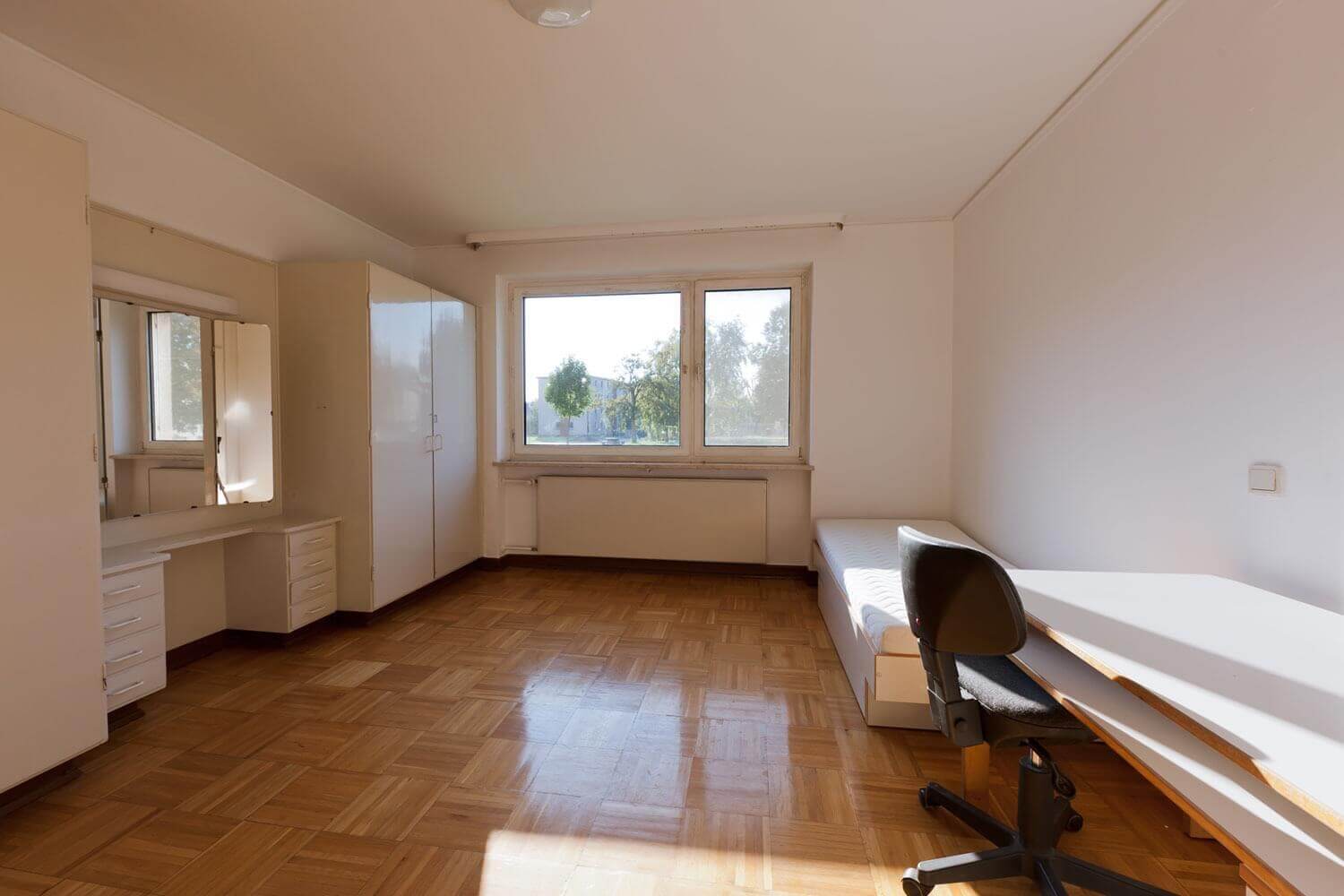 Furnished single room with white closet, integrated chest of drawers with mirror, bed and desk with chair; view through a large window onto green outdoor facilities.