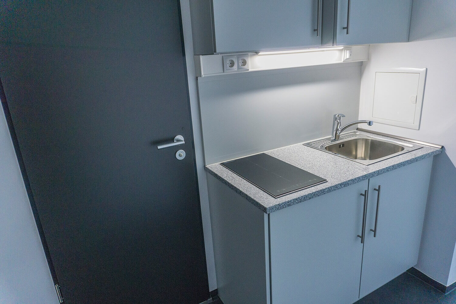 Kleine Küche mit Spülbecken, Zweiplattenkochfeld, Unterschränken, Hängeschränken und dunkler Tür.