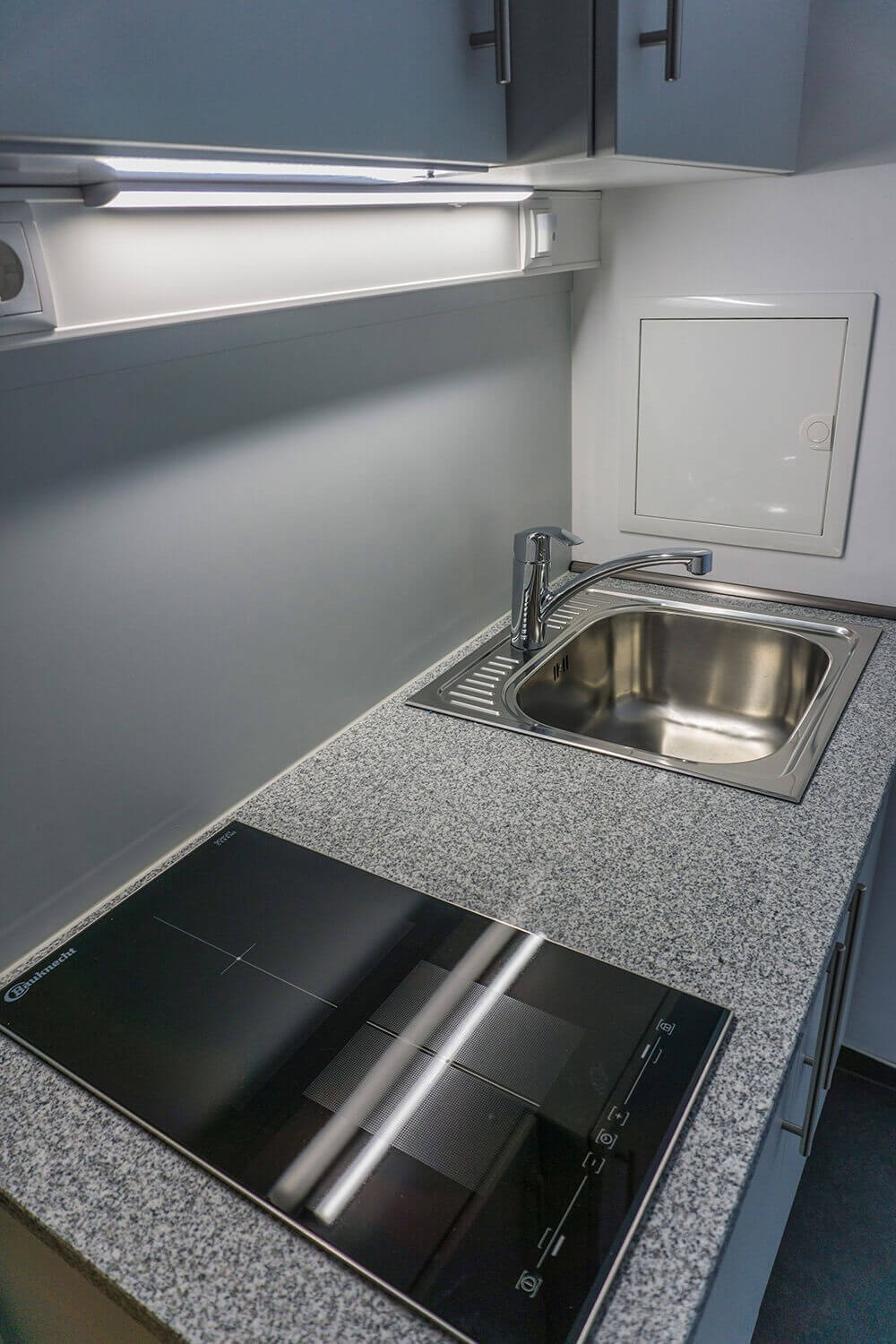 Kompakte Küchenzeile mit grauer Arbeitsplatte, Edelstahlspüle, Cerankochfeld, Hängeschränken und integrierter Beleuchtung unter den Oberschränken.