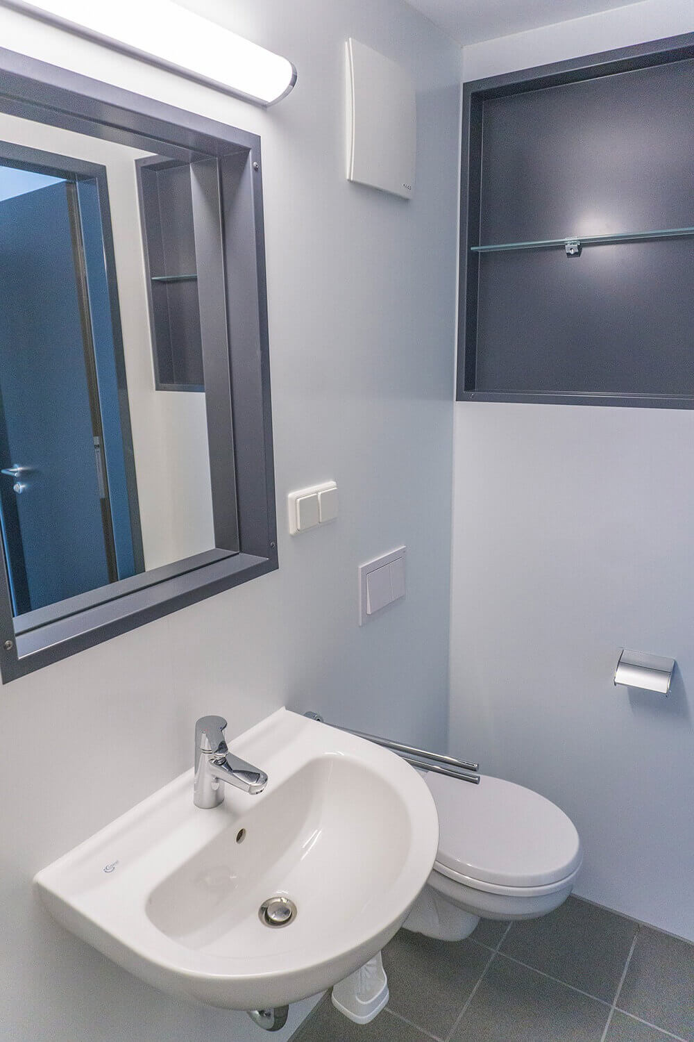 Badezimmer mit wandhängender Toilette, großem Spiegel, Waschbecken, modernem Lichtbalken und Ablageflächen in einem minimalistischen Design.