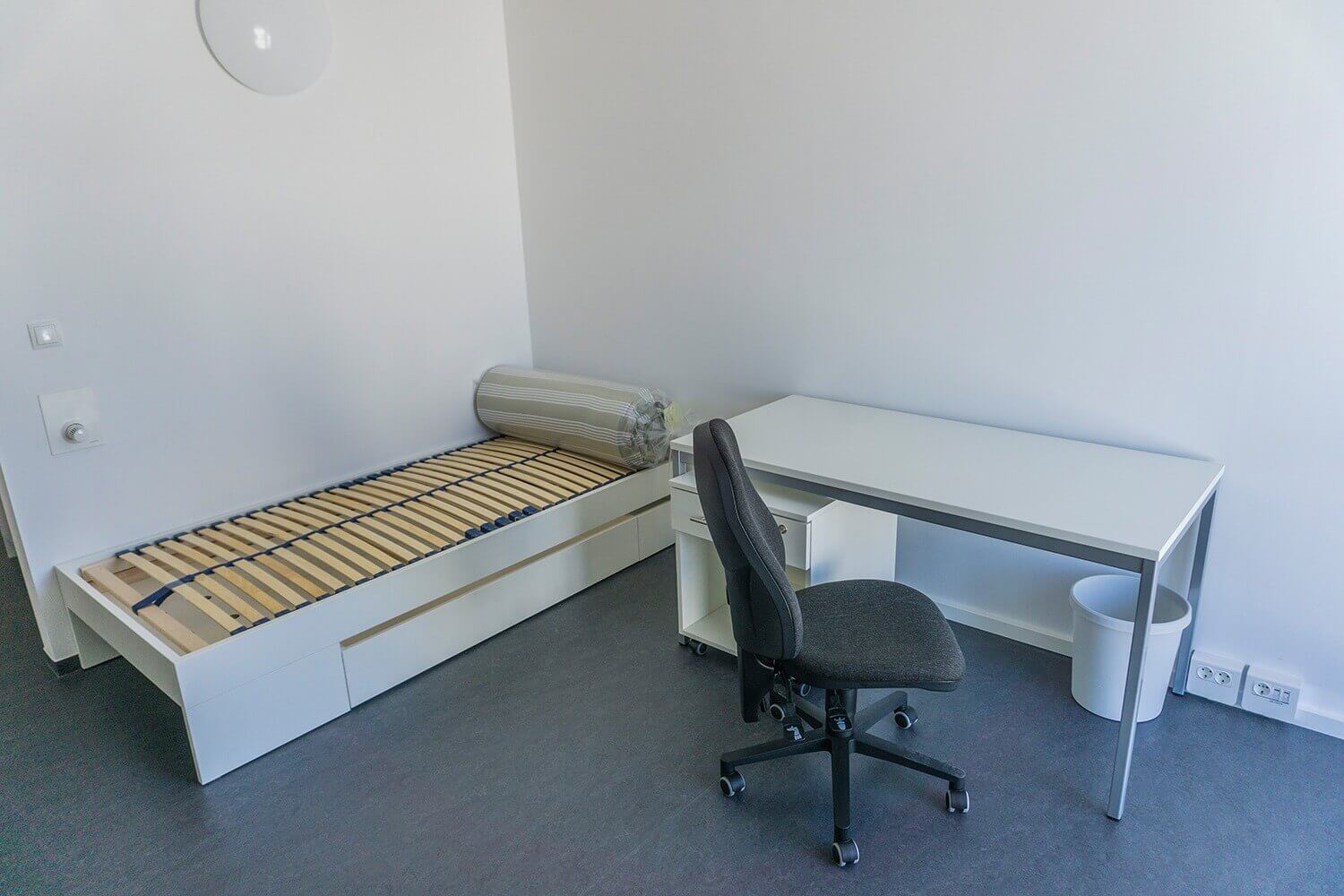 Ein schlicht eingerichtetes Einzelzimmer mit Lattenrostbett, aufgerollter Matratze, weißem Schreibtisch und schwarzem Bürostuhl auf einem dunklen Bodenbelag.