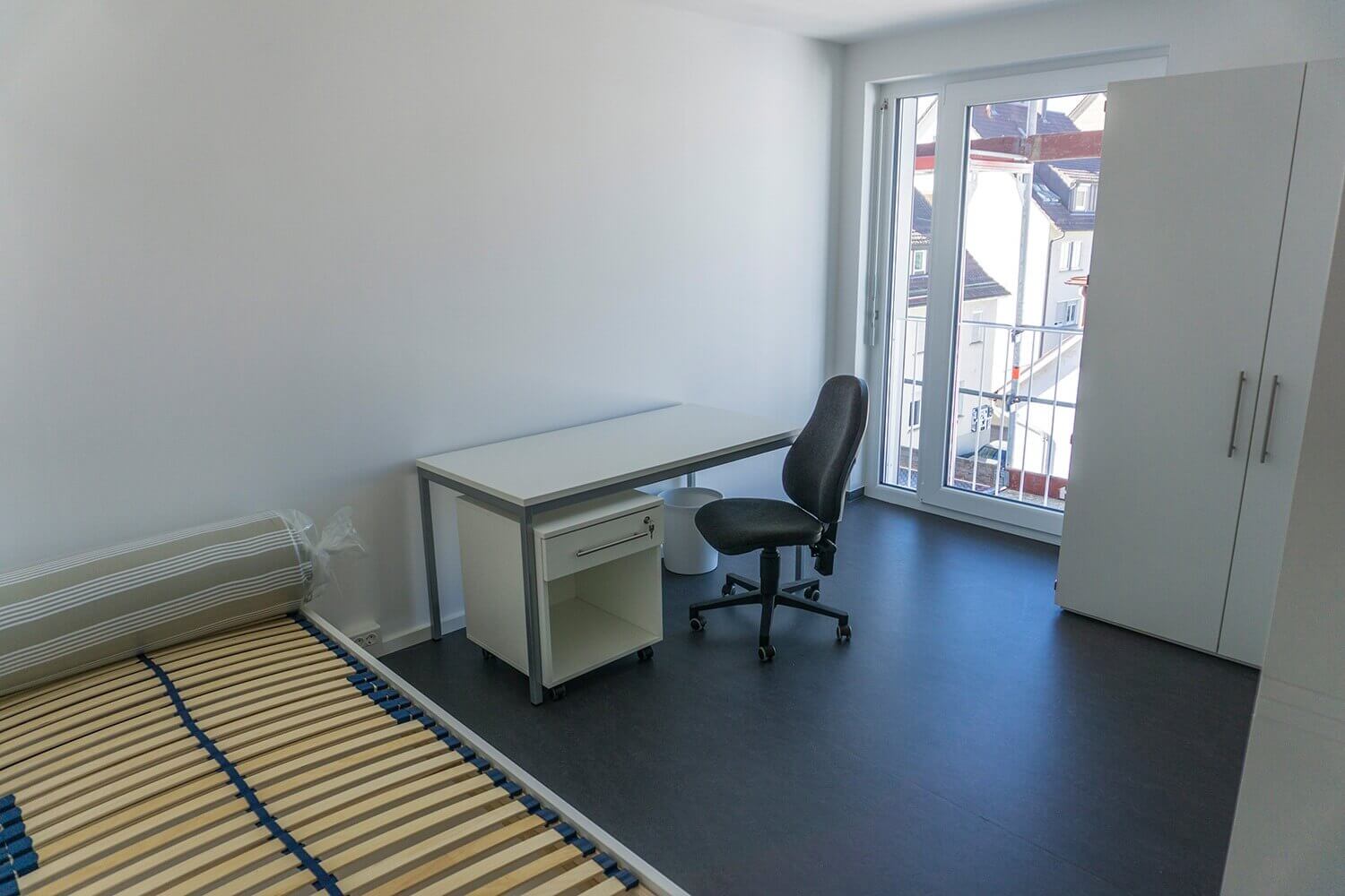 Ein modernes, helles Einzelzimmer mit weißem Schreibtisch, ergonomischem Drehstuhl, großem Fenster mit Blick auf die Nachbarhäuser und einem weißen Kleiderschrank.