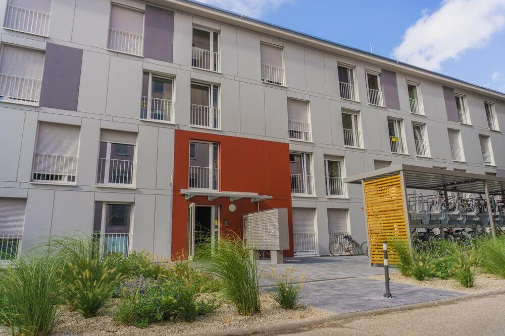 Außenansicht des Wohngebäudes Cäcilienstraße 39 mit rotem Eingangsbereich, gepflegten Grünflächen und einem überdachten Fahrradstellplatz auf der rechten Seite.