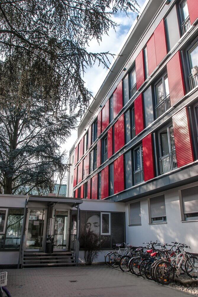 Eingangsbereich des Studentenwohnheims mit Fahrradständern, Rampe, Treppe und Wandbild eines Gesichts.
