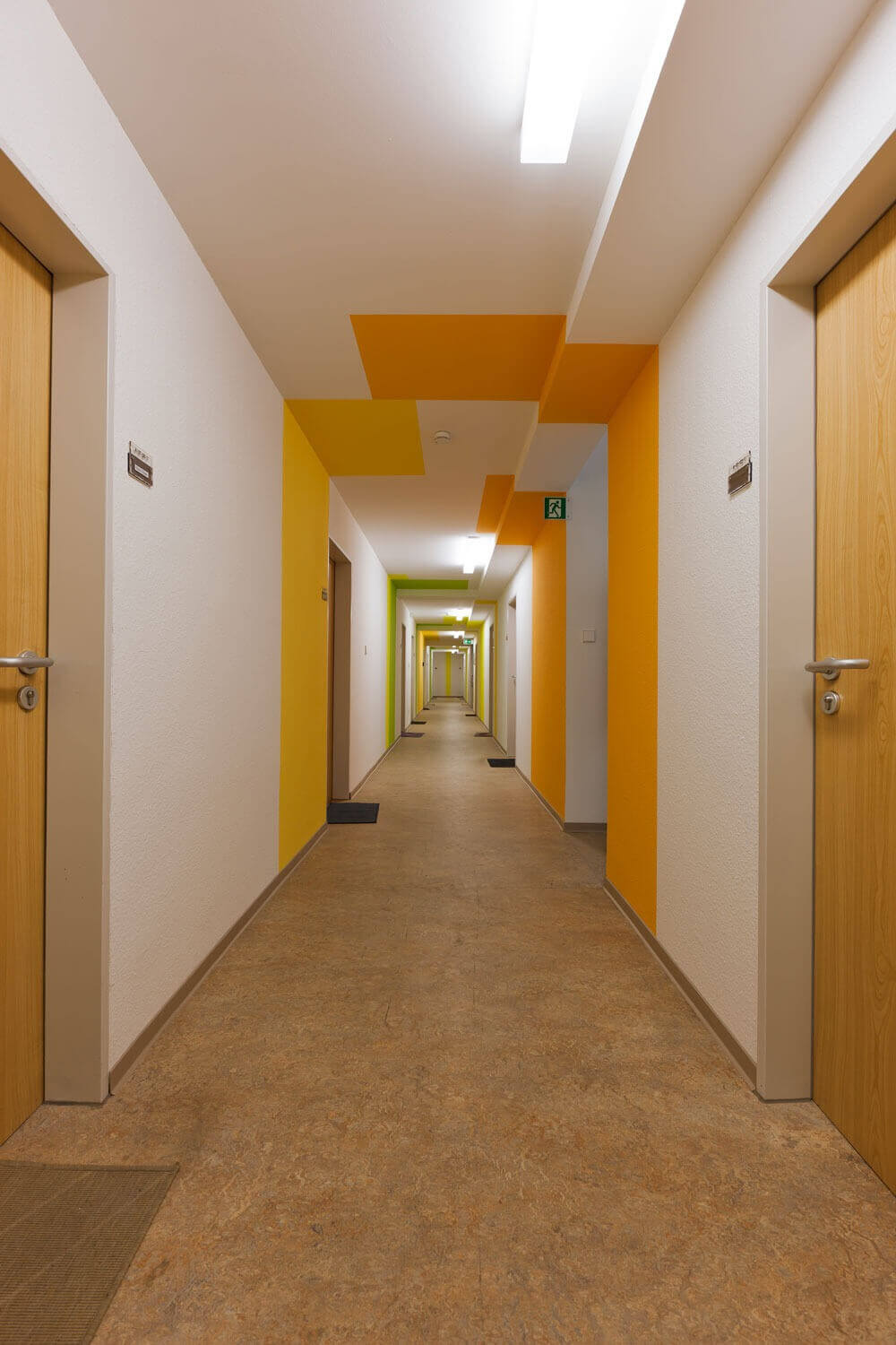 Langer Flur im Studentenwohnheim mit farblich abgesetzten Decken- und Wandbereichen in Gelb- und Orangetönen.