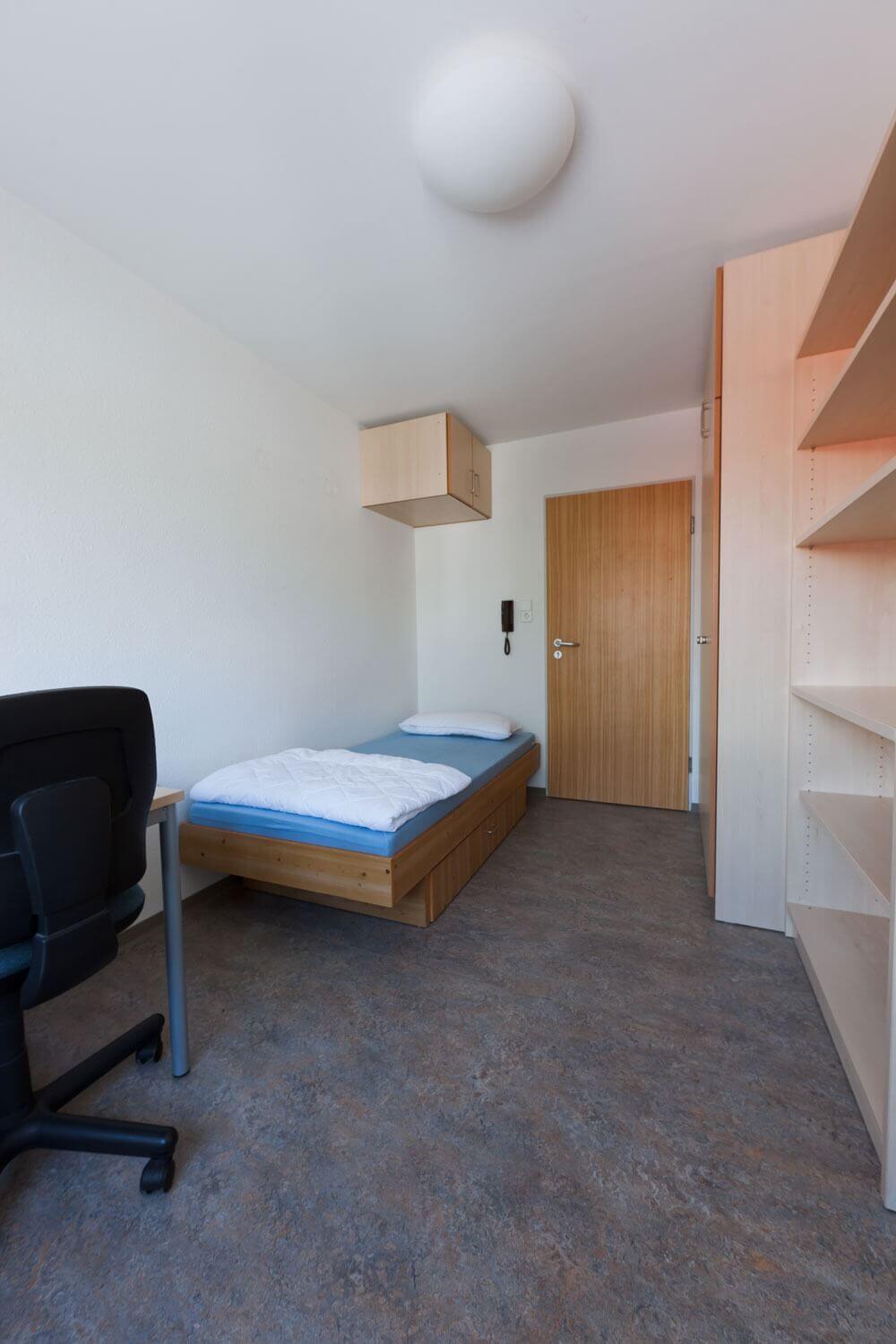 Schlicht eingerichtetes Einzelzimmer im Studentenwohnheim mit Schreibtisch, Regalwand, Bett mit Stauraum und Holztür.