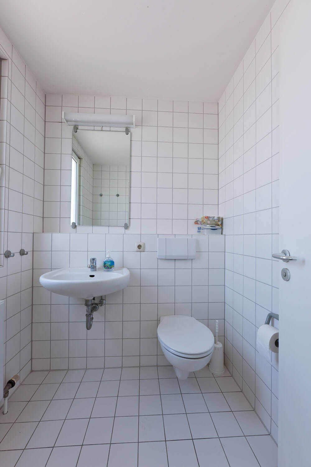 Modern gefliestes Badezimmer mit Tageslicht, Waschbecken, Spiegel, WC und schlichtem, hellen Fliesenspiegel