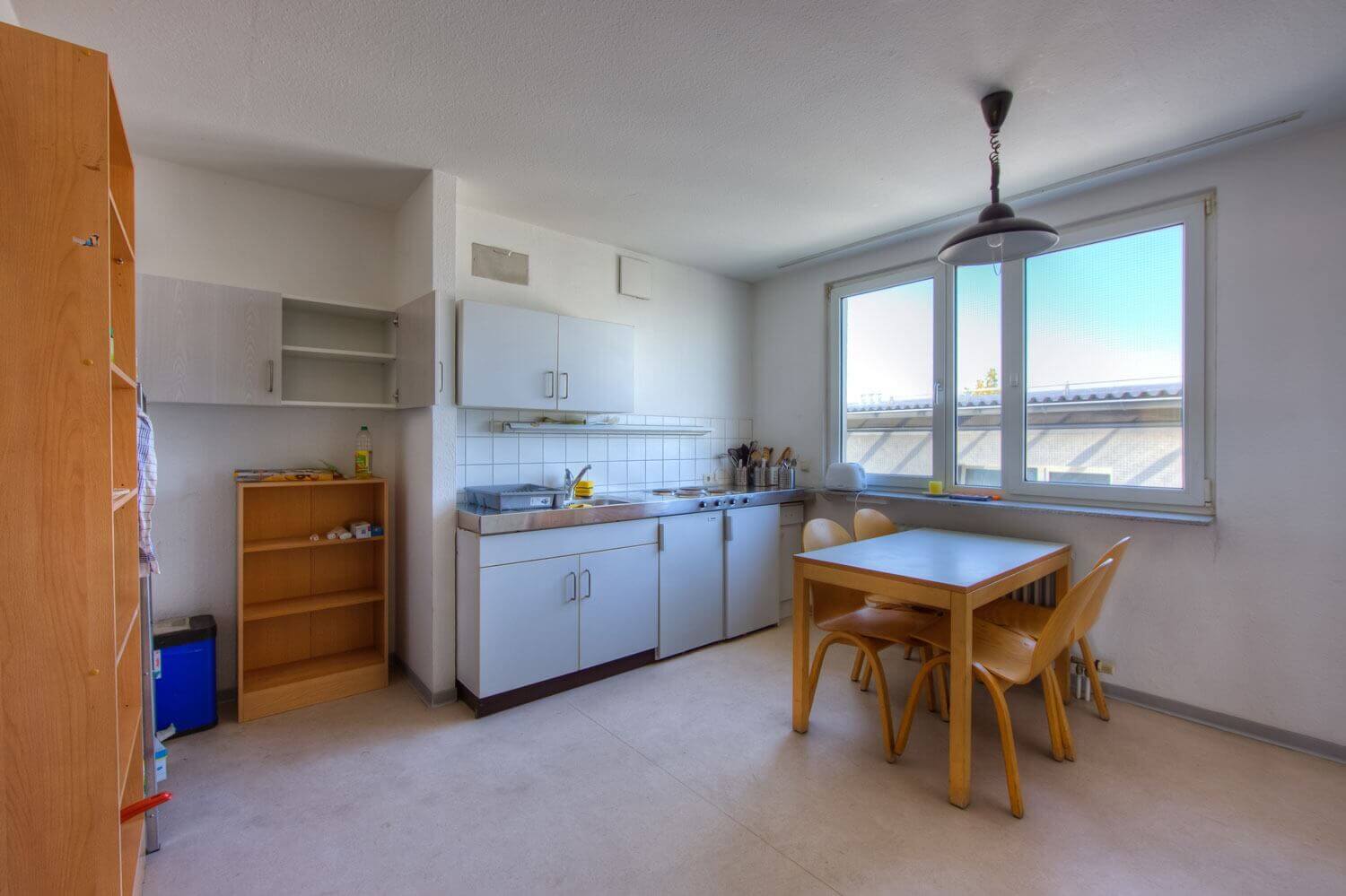 Helle Gemeinschaftsküche mit großem Fenster, Esstisch, vier Stühlen und kompakter Einbauküche mit Hängeschränken