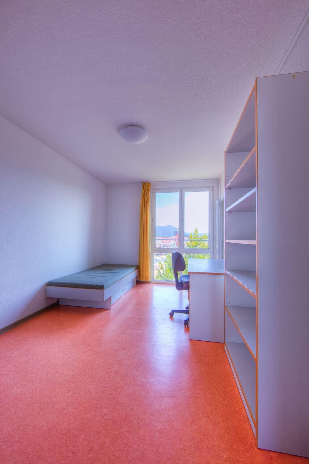 Helles Einzelzimmer mit Bett, Schreibtisch, Stuhl und Regal sowie großem Fenster mit Blick ins Grüne