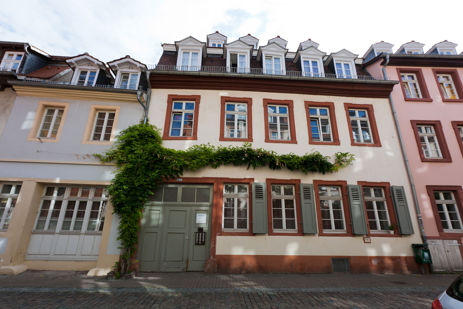 Außenansicht des Europahauses I mit weißen und roten Fensterrahmen, begrüntem Eingangsbereich und Kopfsteinpflaster vor dem Gebäude.