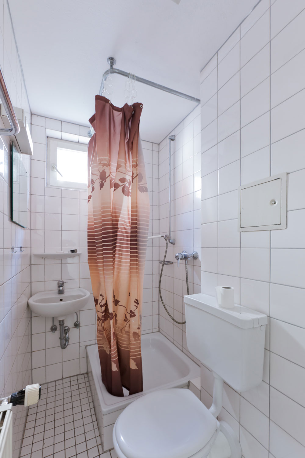 Kleines, funktionales Badezimmer mit Dusche, WC, Waschbecken und Duschvorhang in einer vollständig gefliesten Nische mit Tageslichtfenster.