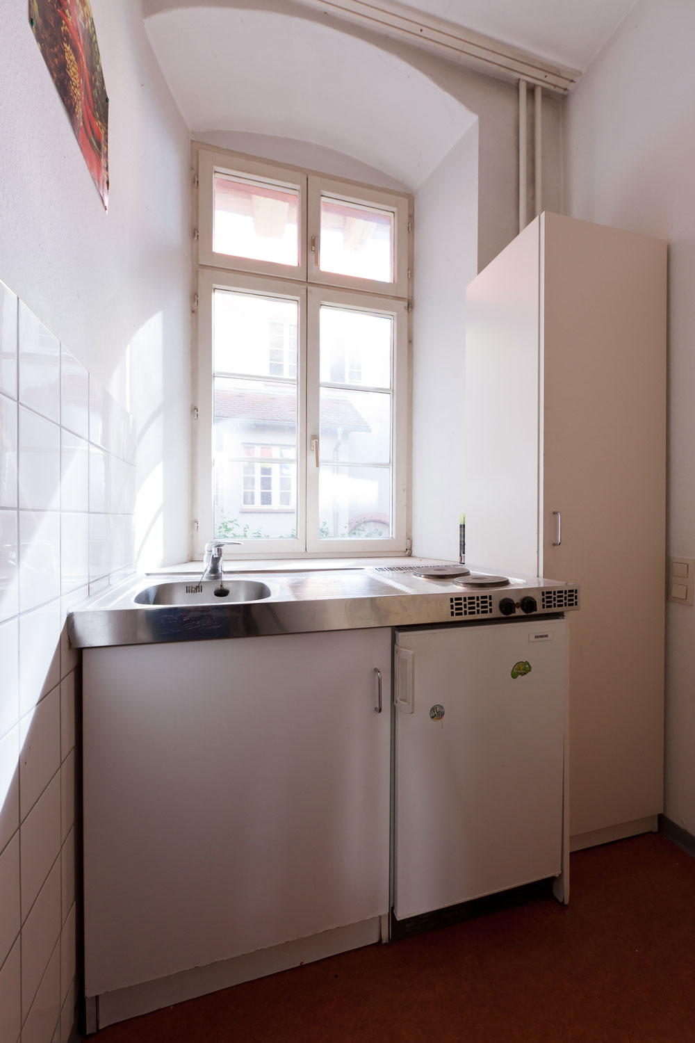 Kompakte Küchenzeile mit Spülbecken, Kochfeld, Kühlschrank und weißem Hängeschrank vor einem großen Fenster mit Blick auf den Innenhof.