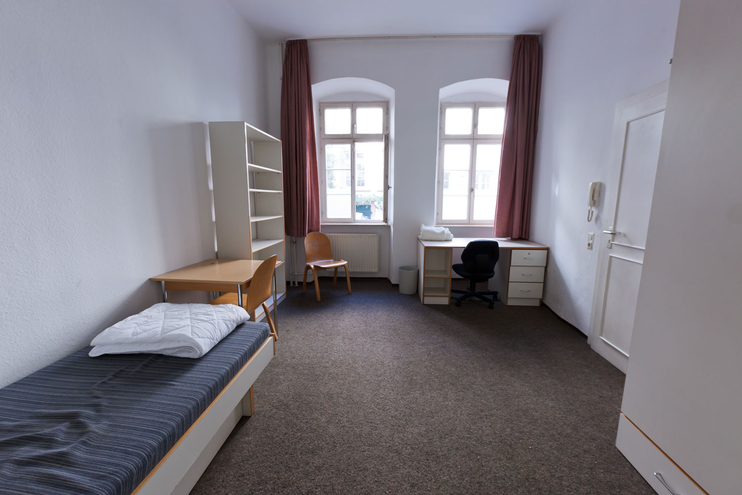 Geräumiges, schlicht möbliertes Einzelzimmer mit Bett, Schreibtisch und einem weiteren Tisch, Bücherregal, Stuhl und Teppichboden, beleuchtet durch zwei große Fenster mit rosa Vorhängen.