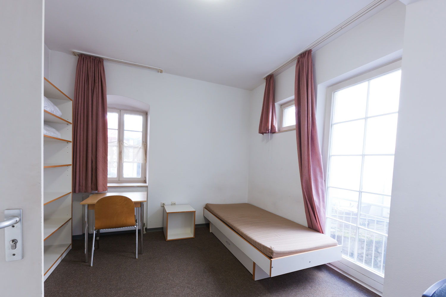 Helles Einzelzimmer mit Bett, Bücherregal, Tisch und zwei Stühlen, ausgestattet mit Teppichboden und großen Fenstern mit rosa Vorhängen.