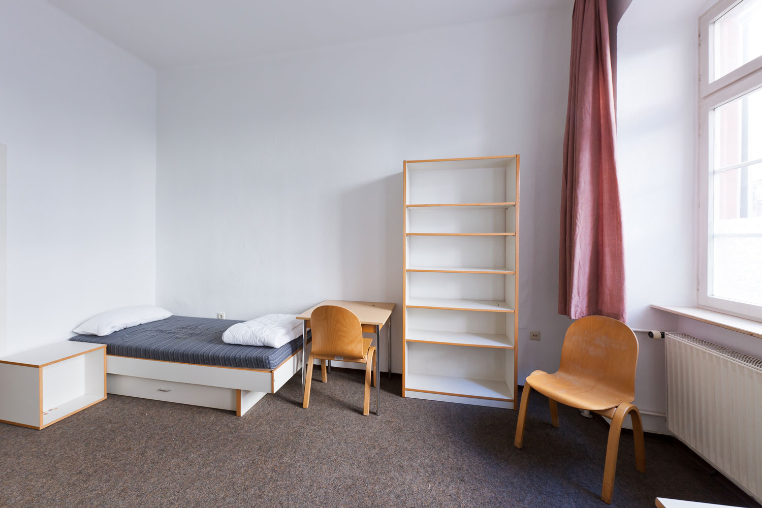 Kleines Wohn- und Schlafzimmer mit Bett, Fensterfront, Vorhängen, Schreibtisch und Regal, eingerichtet für studentisches Wohnen.