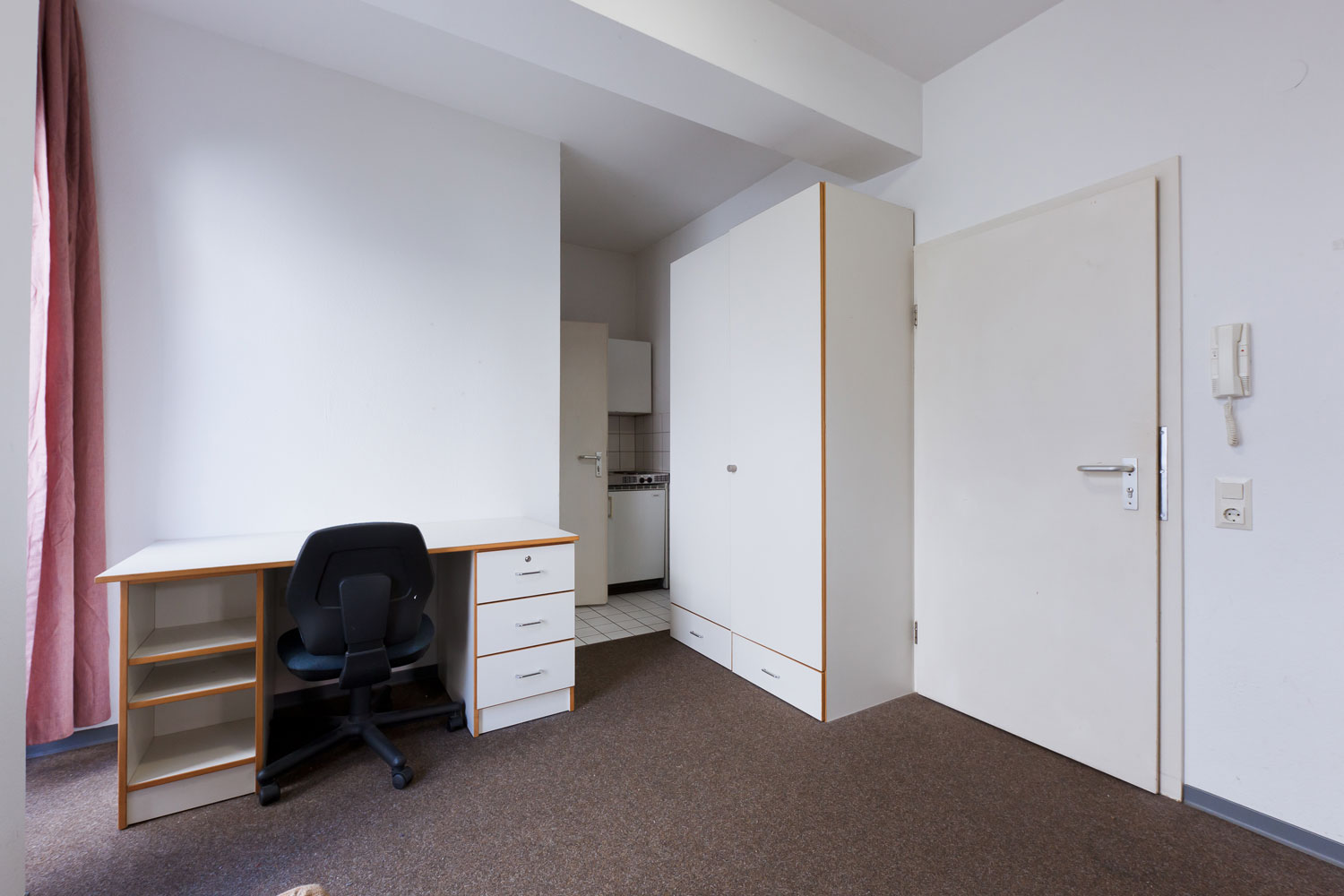 Blick in ein möbliertes Einzelzimmer mit Schreibtisch, Bürostuhl, großem Kleiderschrank und Zugang zur kleinen Pantryküche, Teppichboden und helle Wände runden das funktionale Raumangebot ab.