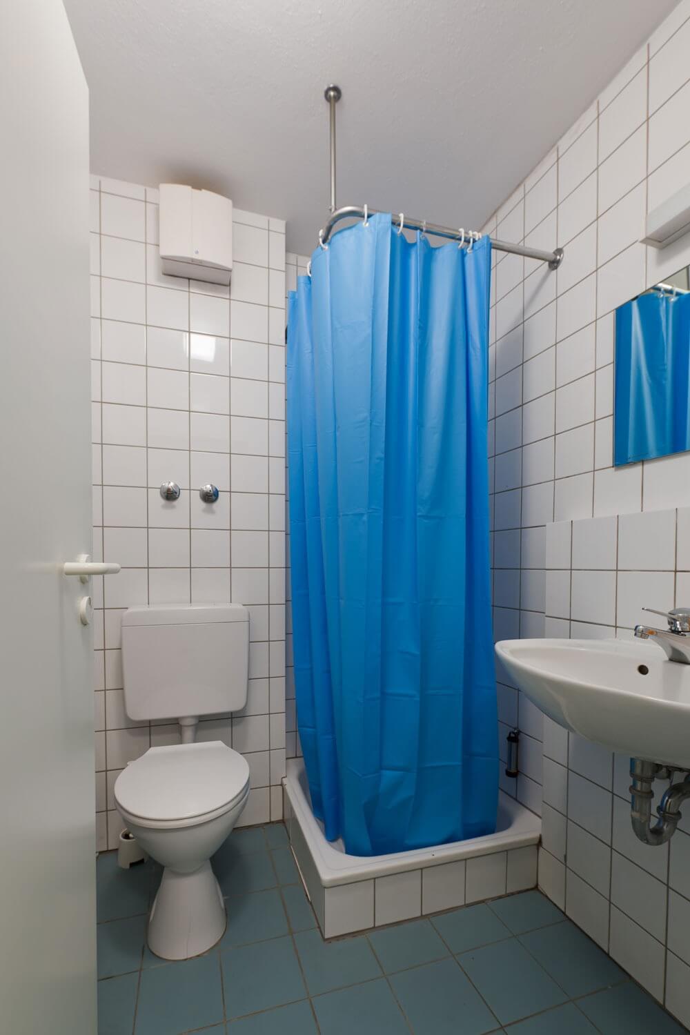 Kompaktes Badezimmer mit Duschkabine mit blauem Duschvorhang, WC und Waschbecken, weiße Fliesen an den Wänden und blaue Fliesen am Boden.