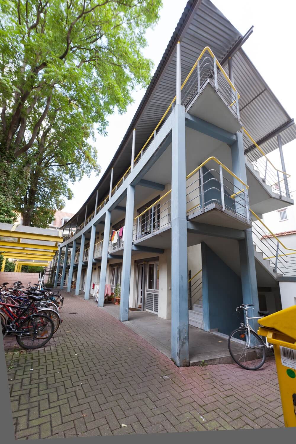 Innenhof eines Wohnhauses mit offener Laubengangerschließung, außenliegenden Treppenaufgängen, blaugrauen Stützen und gelben Geländern, flankiert von Fahrradstellplätzen und begrüntem Baumbestand.