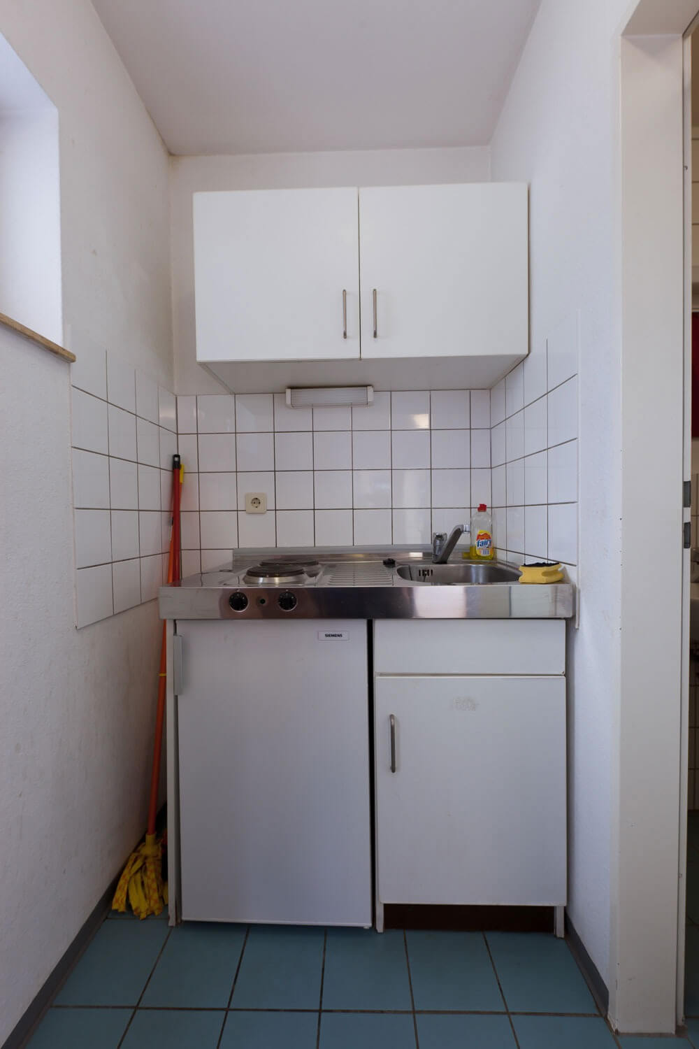 Kompakte Kochnische mit weißem Unterschrank, kleinem Kühlschrank, Spüle und zwei Herdplatten, umrahmt von weiß gefliesten Wänden.