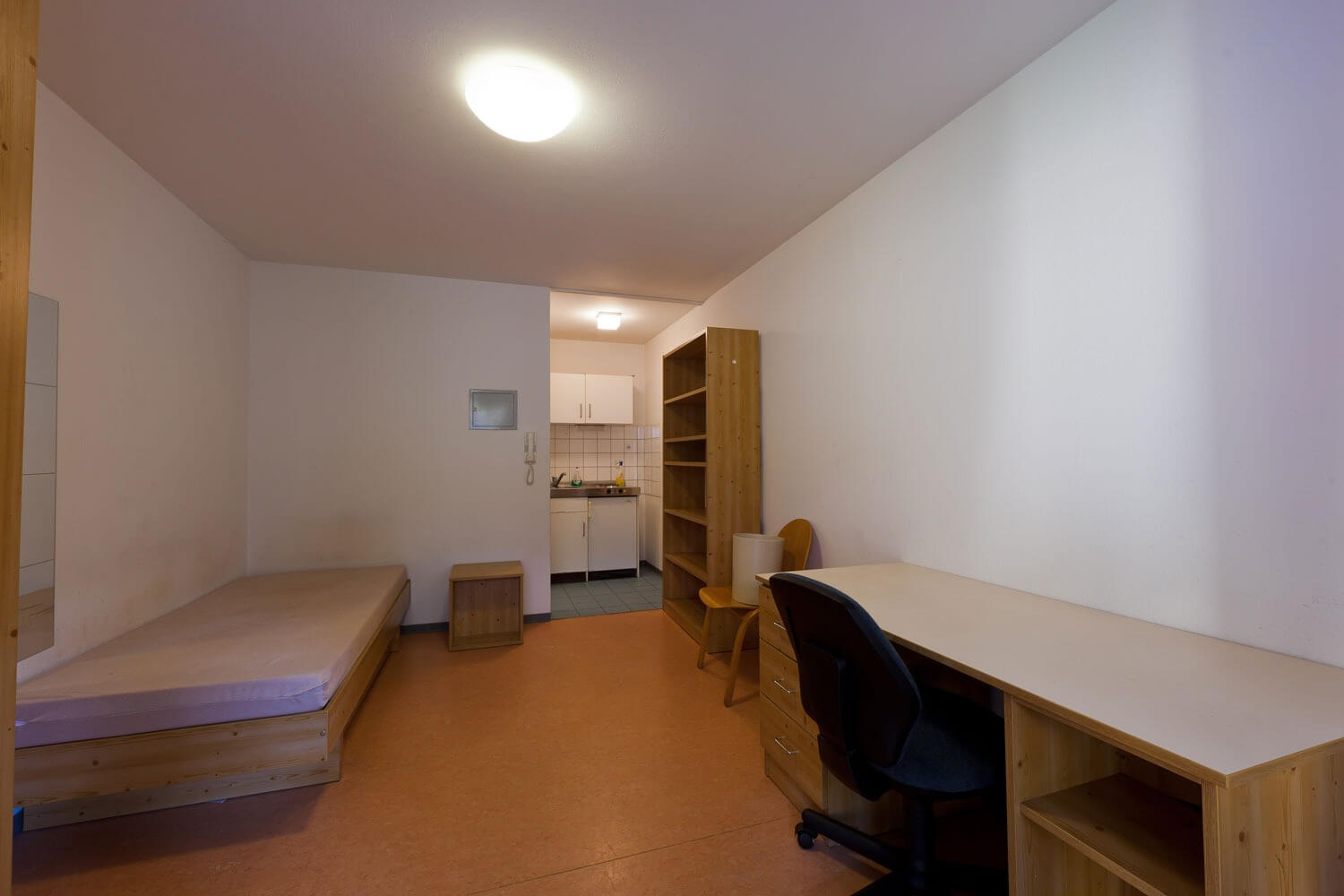 Blick in ein funktional eingerichtetes Zimmer mit Bett, Nachttisch, großem Schreibtisch und Regal aus Holz, im Hintergrund eine kleine Kochnische.