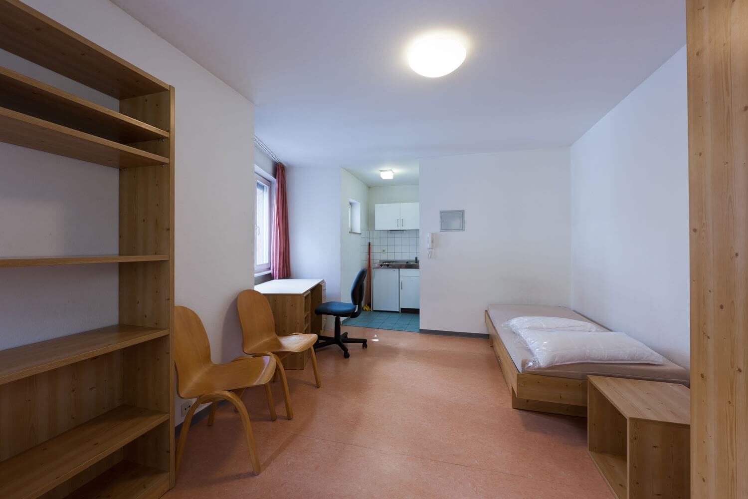 Einrichtung eines Studentenzimmers mit Schreibtisch, zwei Holzstühlen, Regal und Einzelbett mit Kissen, dahinter eine kleine Küchenzeile mit Fliesenboden.