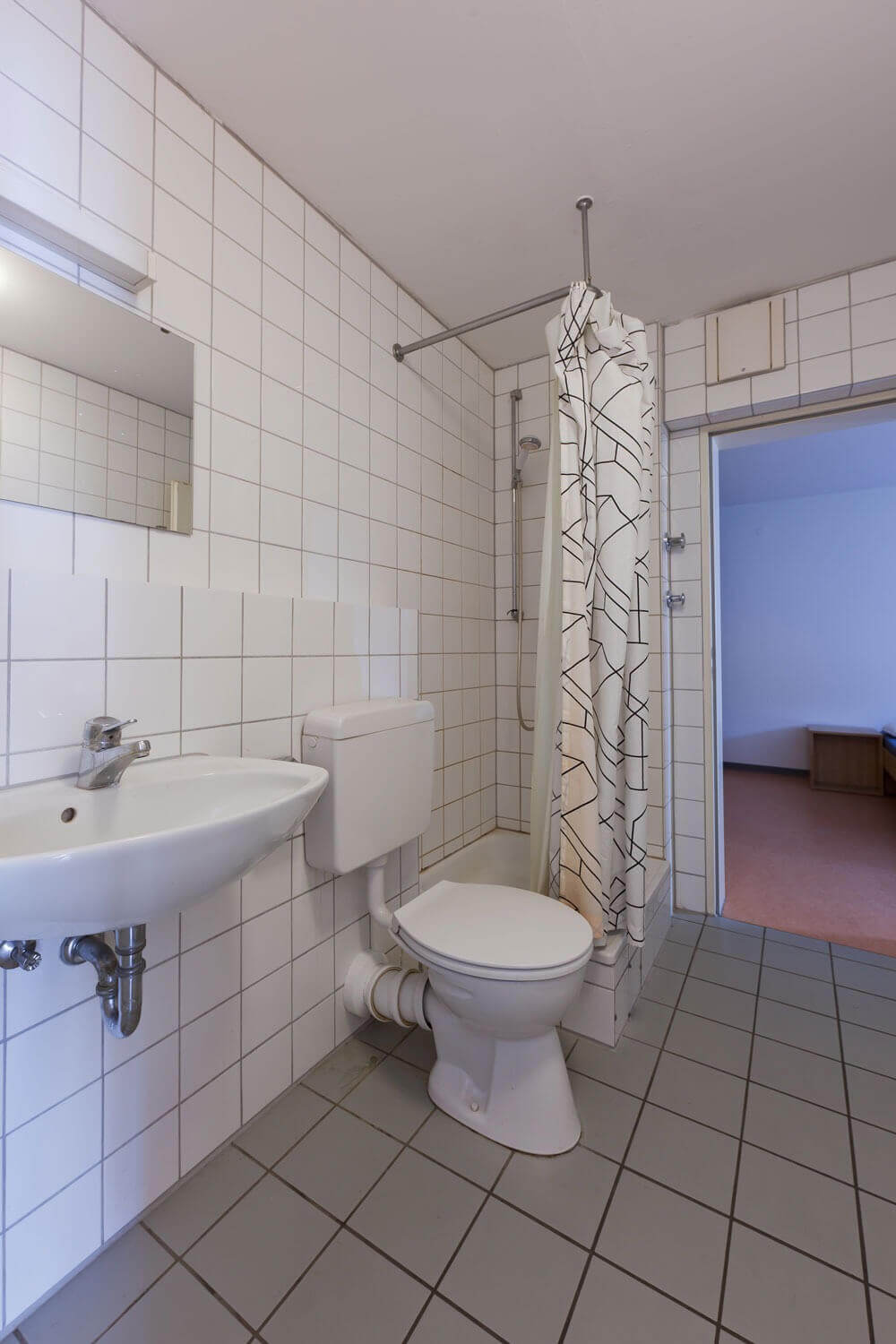 Sanitärbereich mit weiß gefliesten Wänden, Toilette, Waschbecken, Spiegel und einer Duschkabine mit Duschvorhang in Schwarz-Weiß-Muster.