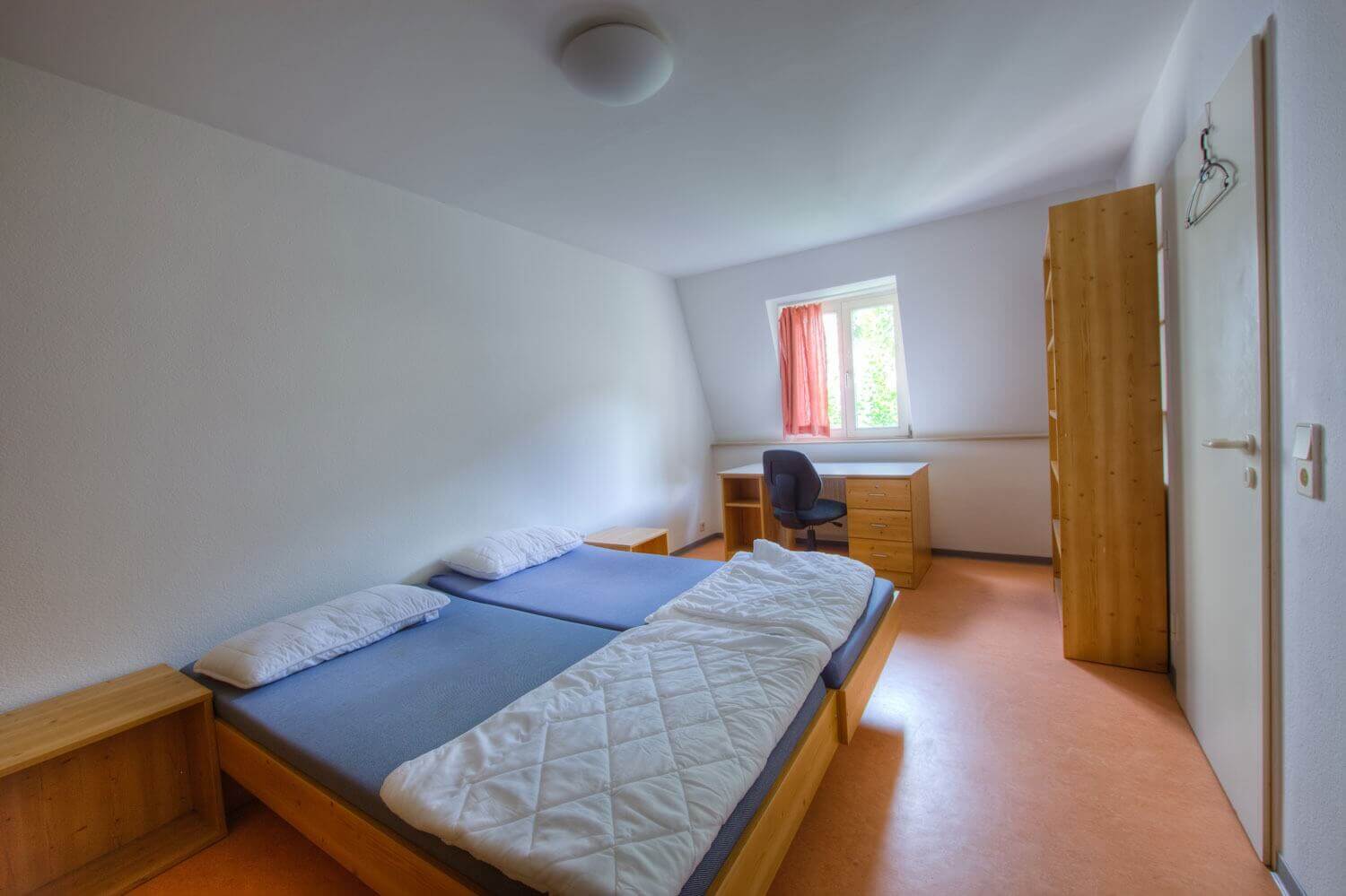 Helles Zimmer mit zwei nebeneinander stehenden Betten, Nachttischen, einem großen Schreibtisch mit Schubladen und Blick auf ein Fenster mit Vorhängen.