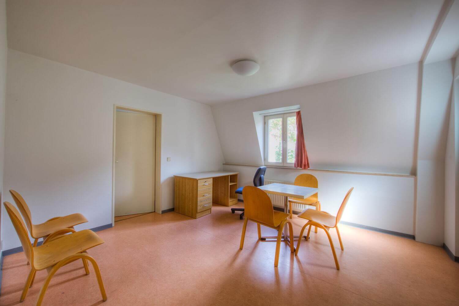 Großer Raum mit mehreren Holzstühlen, einem kleinen Tisch, einem Schreibtisch und einem Fenster mit roten Vorhängen, das Tageslicht hereinlässt.