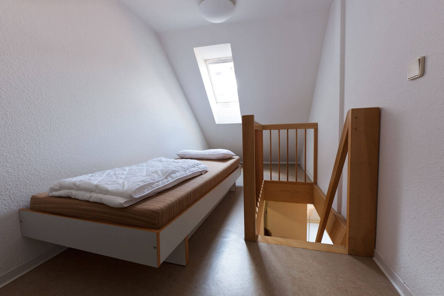 Kleines Dachzimmer mit schräger Wand und Dachfenster, ausgestattet mit einem Einzelbett und einem Geländer, das die Treppe nach unten sichert.