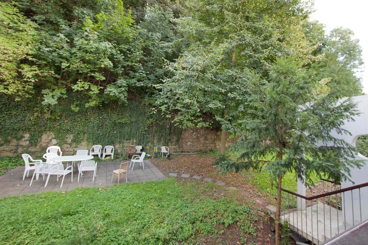 Grünfläche hinter dem Europahaus 5 mit Sitzbereich aus weißen Kunststoffstühlen und Tischen unter schattenspendenden Bäumen.