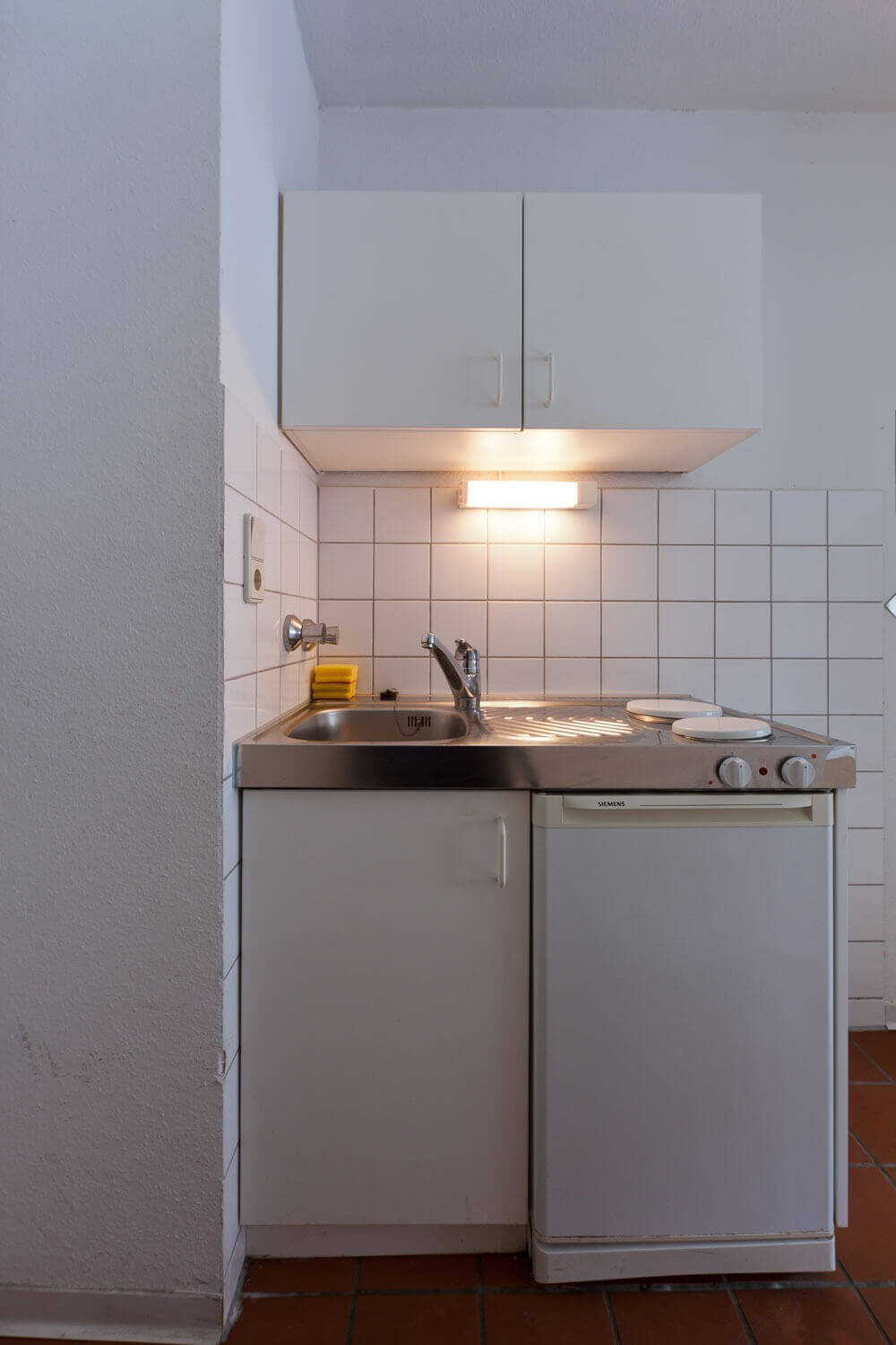 Kompakte Kochnische im Europahaus 5 mit weißem Fliesenspiegel, Spüle, zwei Herdplatten, Unterschrank und Hängeschrank.