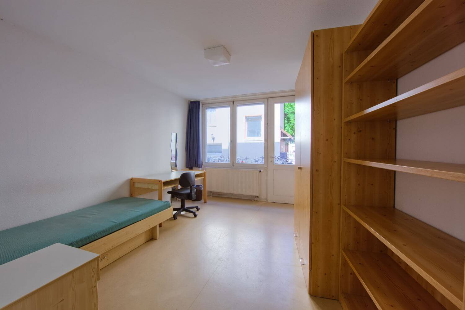 Helles Einzelzimmer im Europahaus 5 mit großem Fenster, Schreibtisch, Drehstuhl, Wandregal, Kleiderschrank und Blick auf den Innenhof.