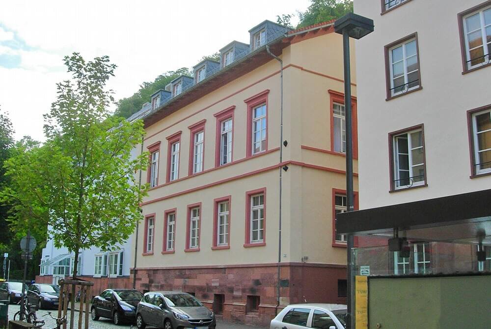 Straßenansicht des Fremeryhauses mit hellbeiger Fassade, roten Fensterrahmen und angrenzenden Stadtgebäuden.