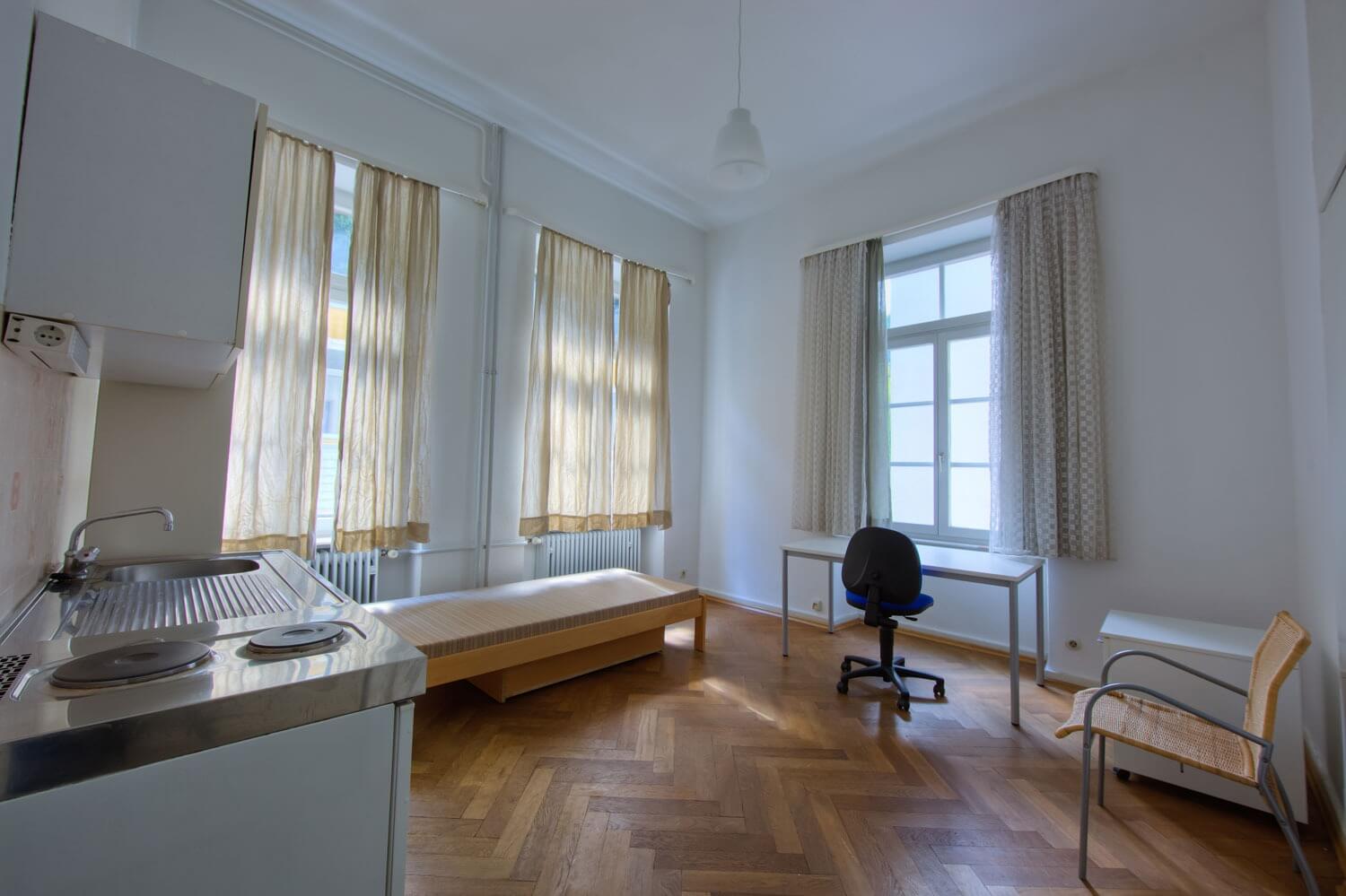 Ein möbliertes Einzelappartement mit Küchenzeile, Bett, Schreibtisch und Parkettboden vor großen Fenstern mit Vorhängen.