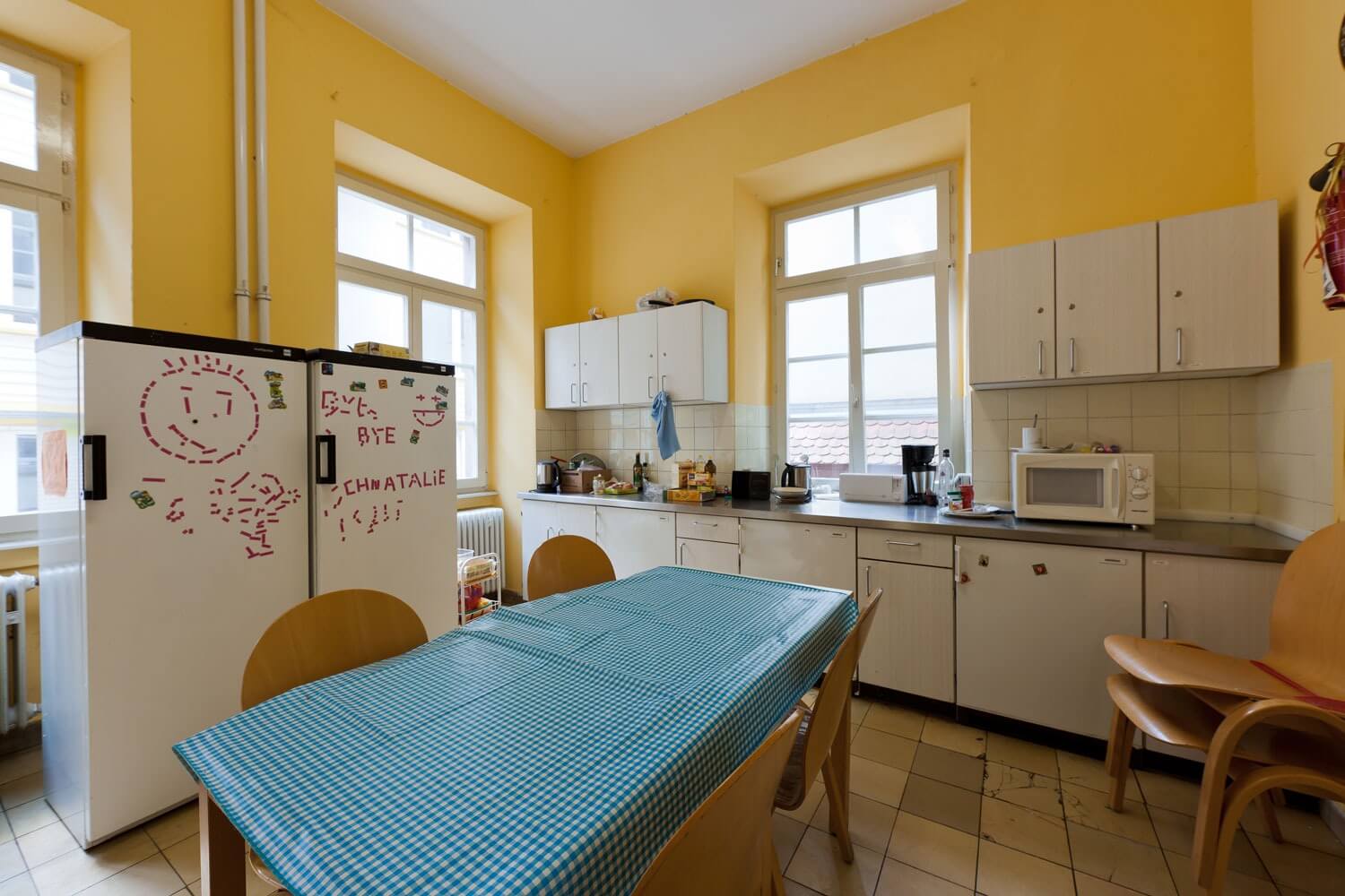 Große Gemeinschaftsküche mit gelben Wänden, Tisch mit Wachstuchtischdecke, weißen Hängeschränken und beschrifteten Kühlschränken.