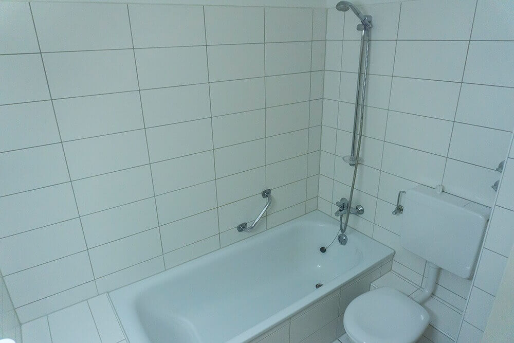 Einfaches Badezimmer mit Badewanne, Duschkopf, WC und weißen Wandfliesen in einem Studentenwohnheim.