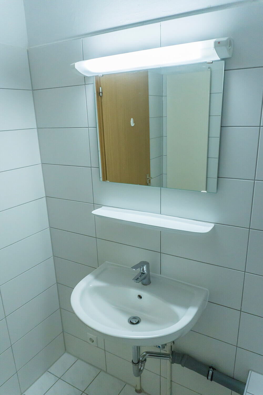 Weiß gefliestes Badezimmer mit Spiegel, Waschbecken und schmalem Regal in einem Studentenwohnheim.