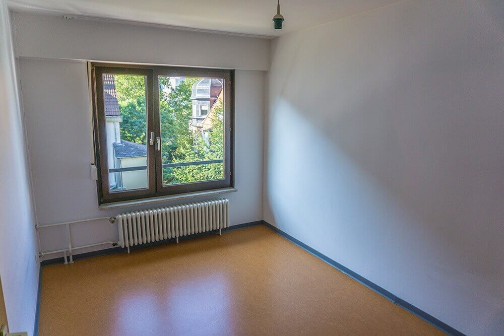 Schlichtes Einzelzimmer mit großem Fenster, Heizkörper und hellem Linoleumboden in einem Studentenwohnheim.