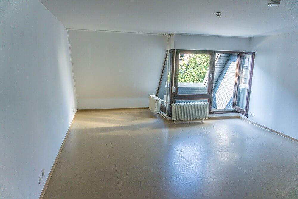 Leeres, helles Zimmer im Studentenwohnheim mit großem Fenster, Heizkörper und Zugang zu einem kleinen Balkonbereich unter dem Dach.