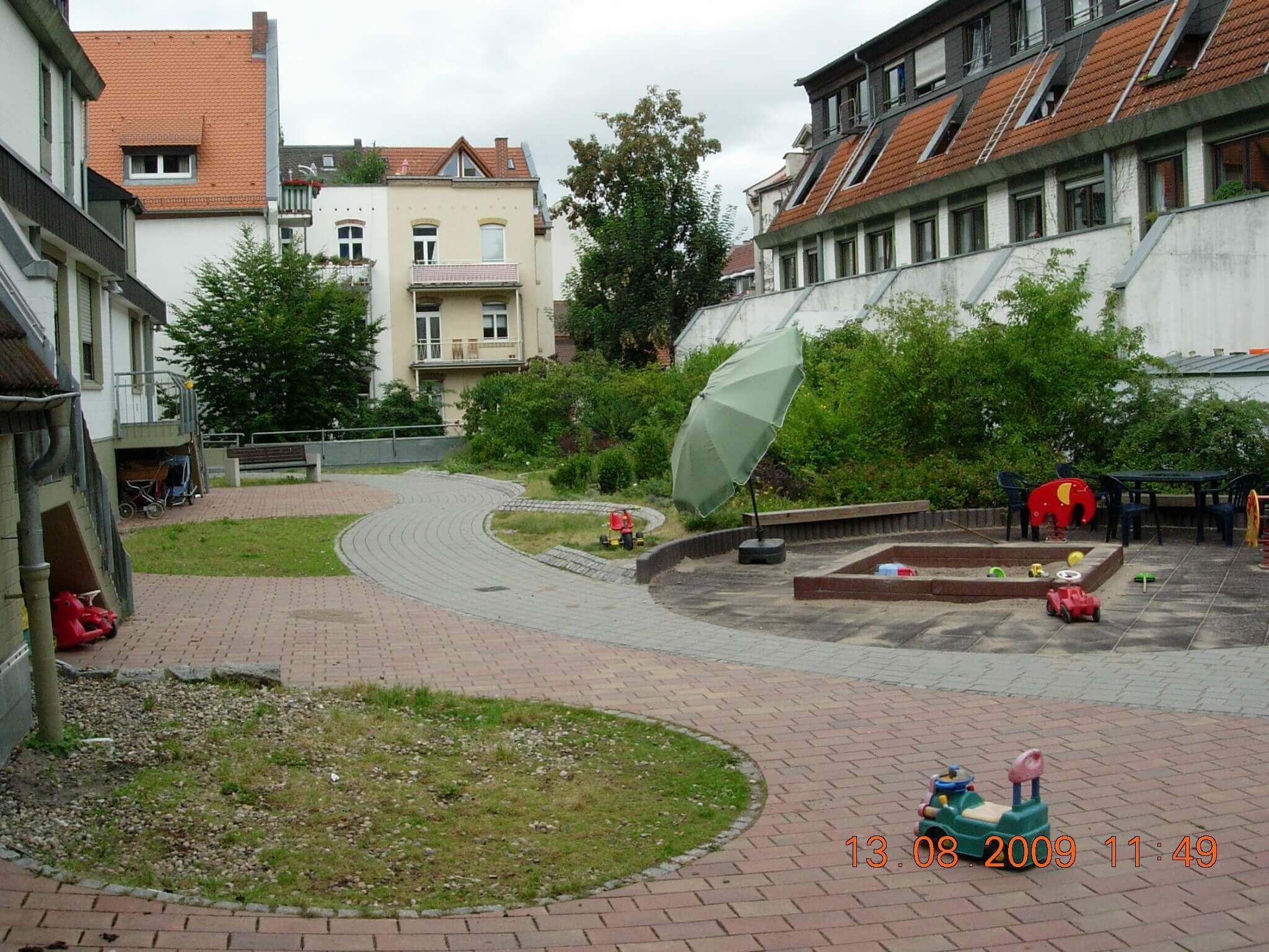Gepflasterter Innenhof eines Studentenwohnheims mit Spielplatz, Sandkasten, Spielzeug und Sonnenschirm, umgeben von Gebäuden mit Satteldächern und Balkonen.